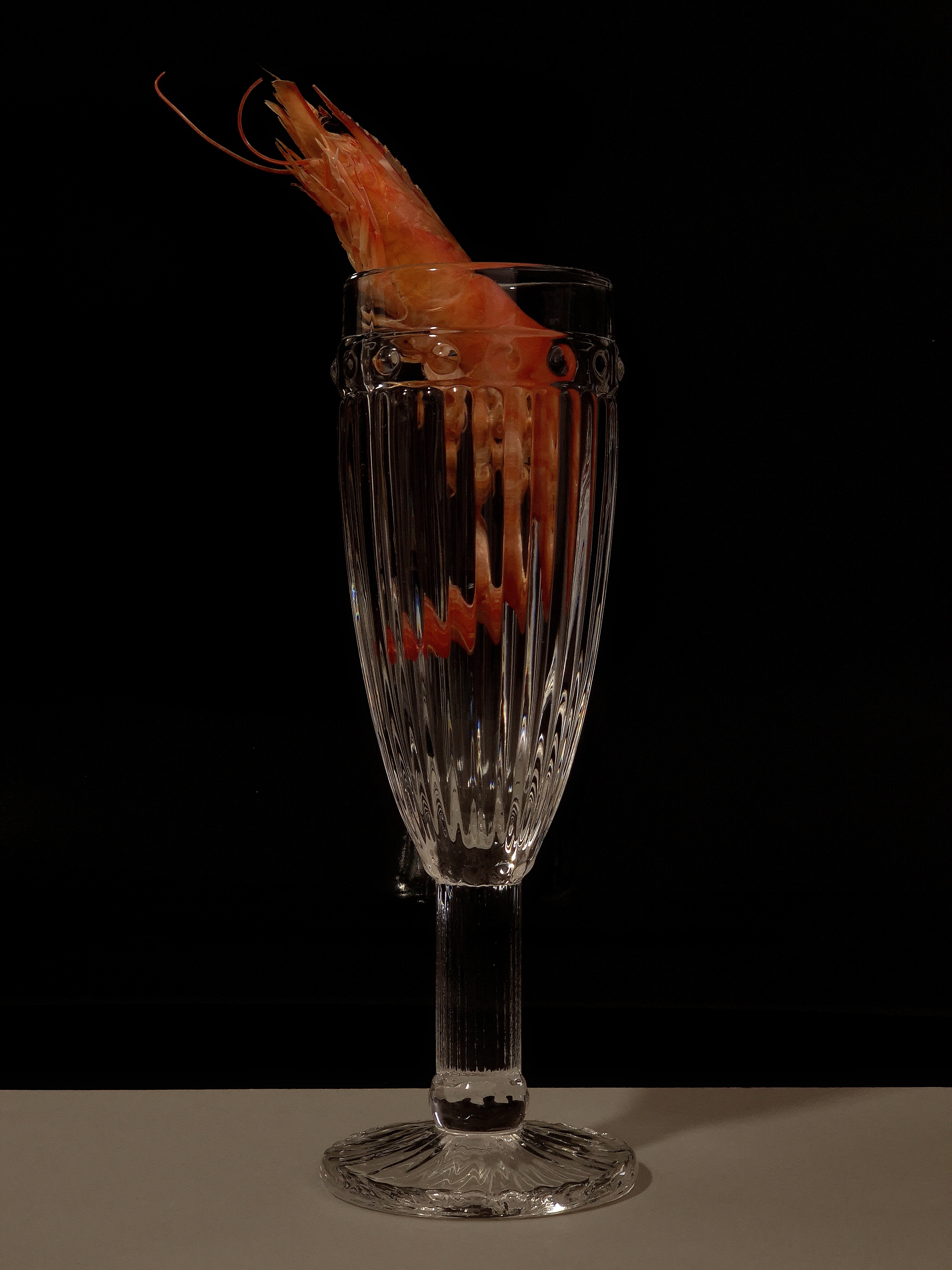 Still life of a shrimp placed inside a tall cut-crystal glass against a dark background.