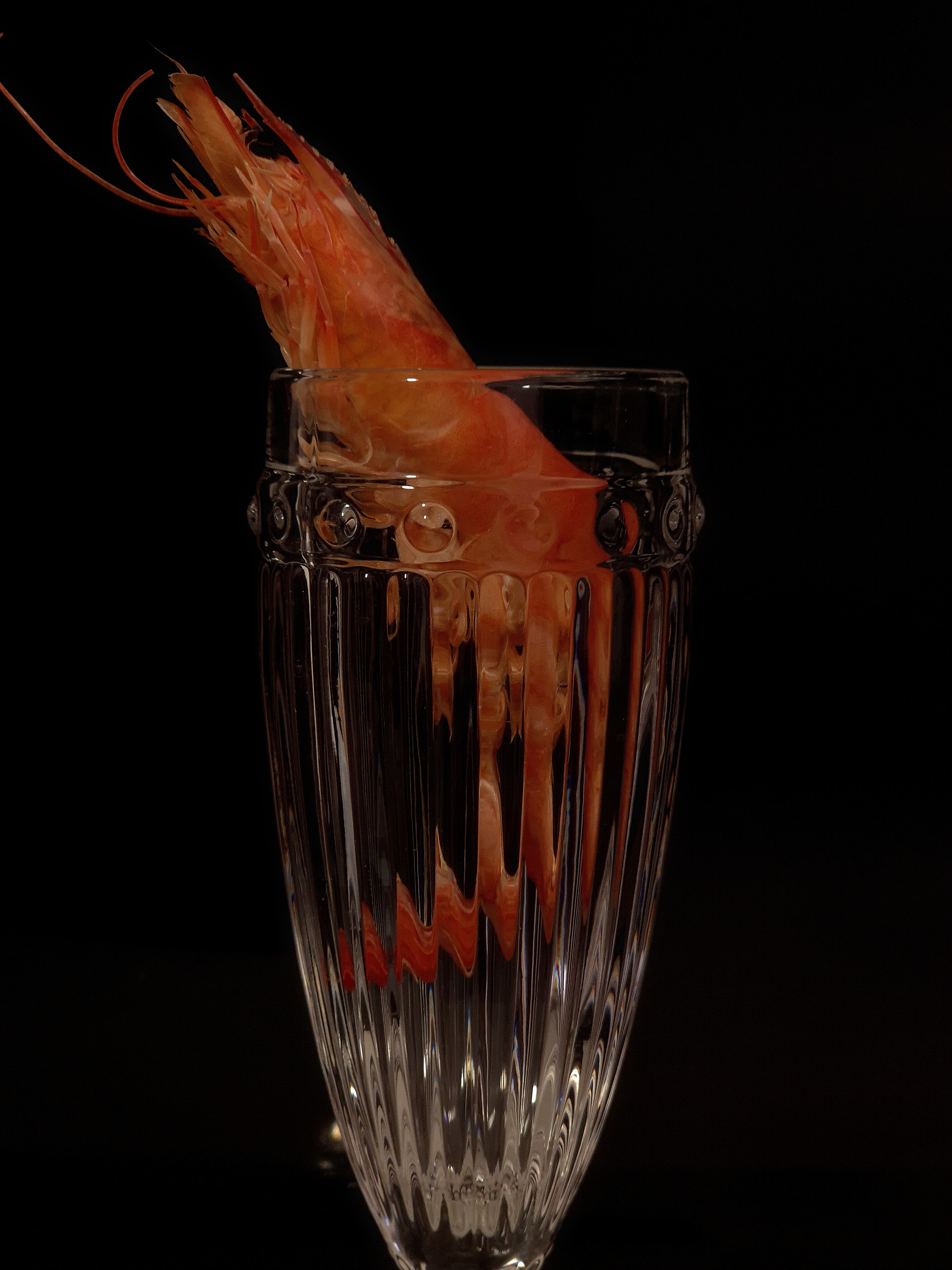 Close-up still life of a shrimp emerging from a tall cut-crystal glass against a black background.