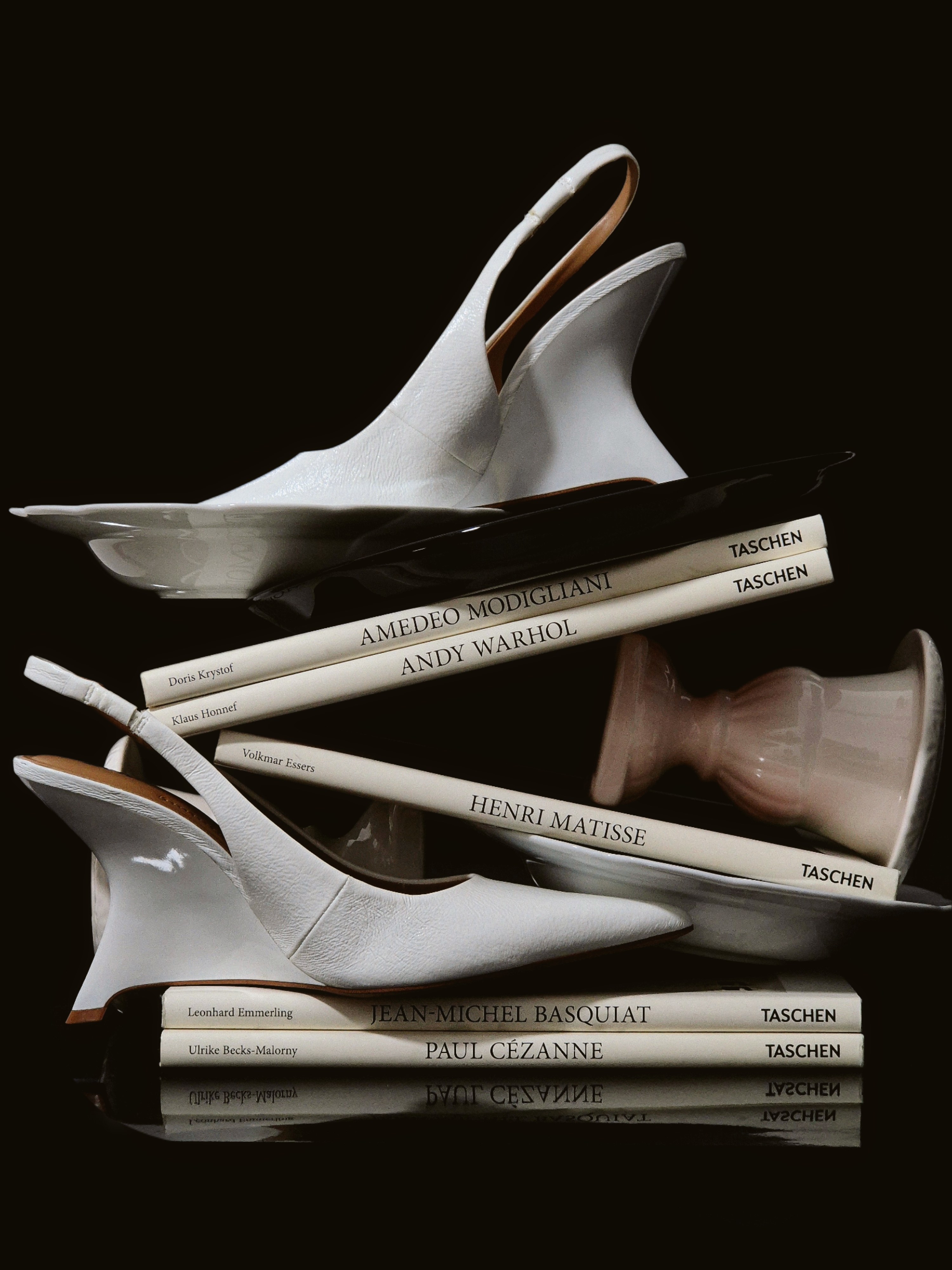 White sculptural heels arranged over stacked art books and porcelain pieces on a black background in a refined still life composition.