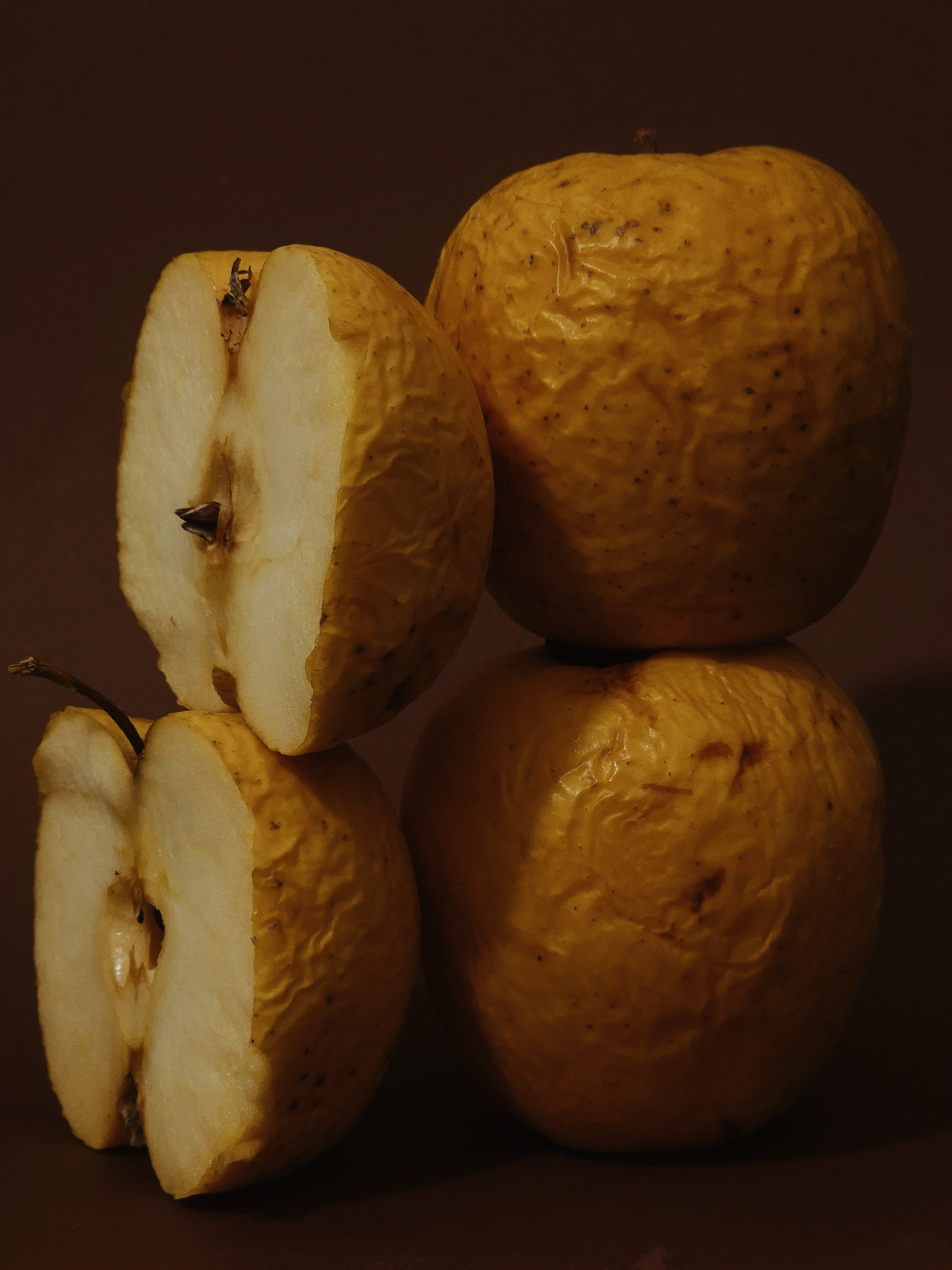 Still life of wrinkled yellow apples stacked against a brown backdrop.