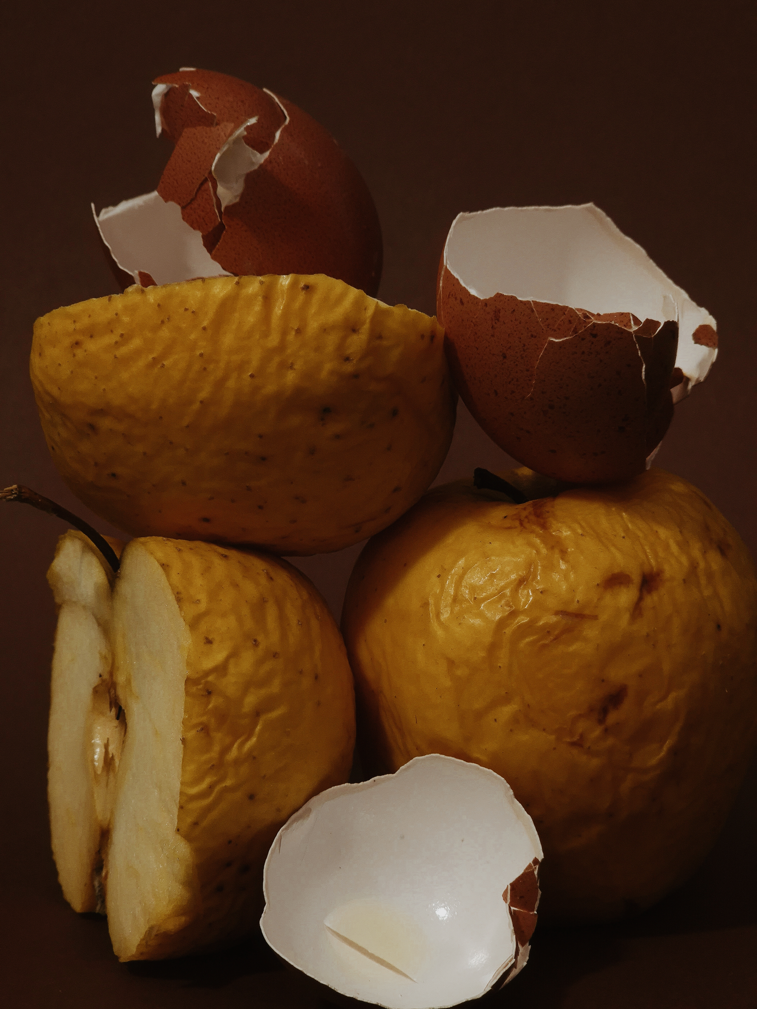 Still life featuring bruised apples and broken eggshells arranged in a sculptural composition against a brown background.