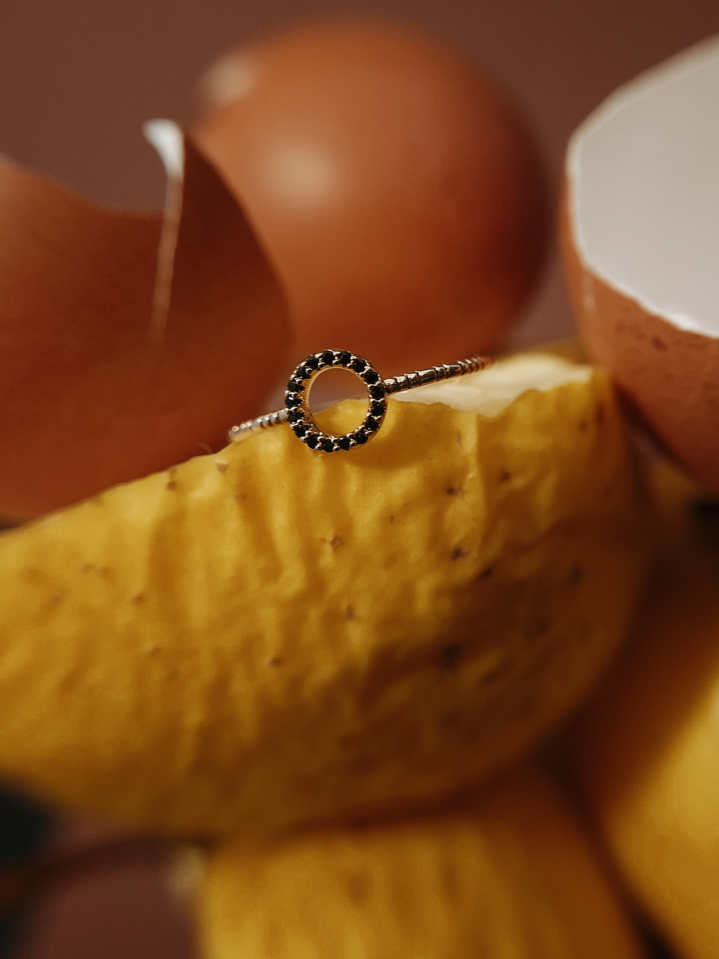 Macro close-up of a delicate gold ring resting on the surface of a wrinkled yellow apple with blurred eggshells in the background.
