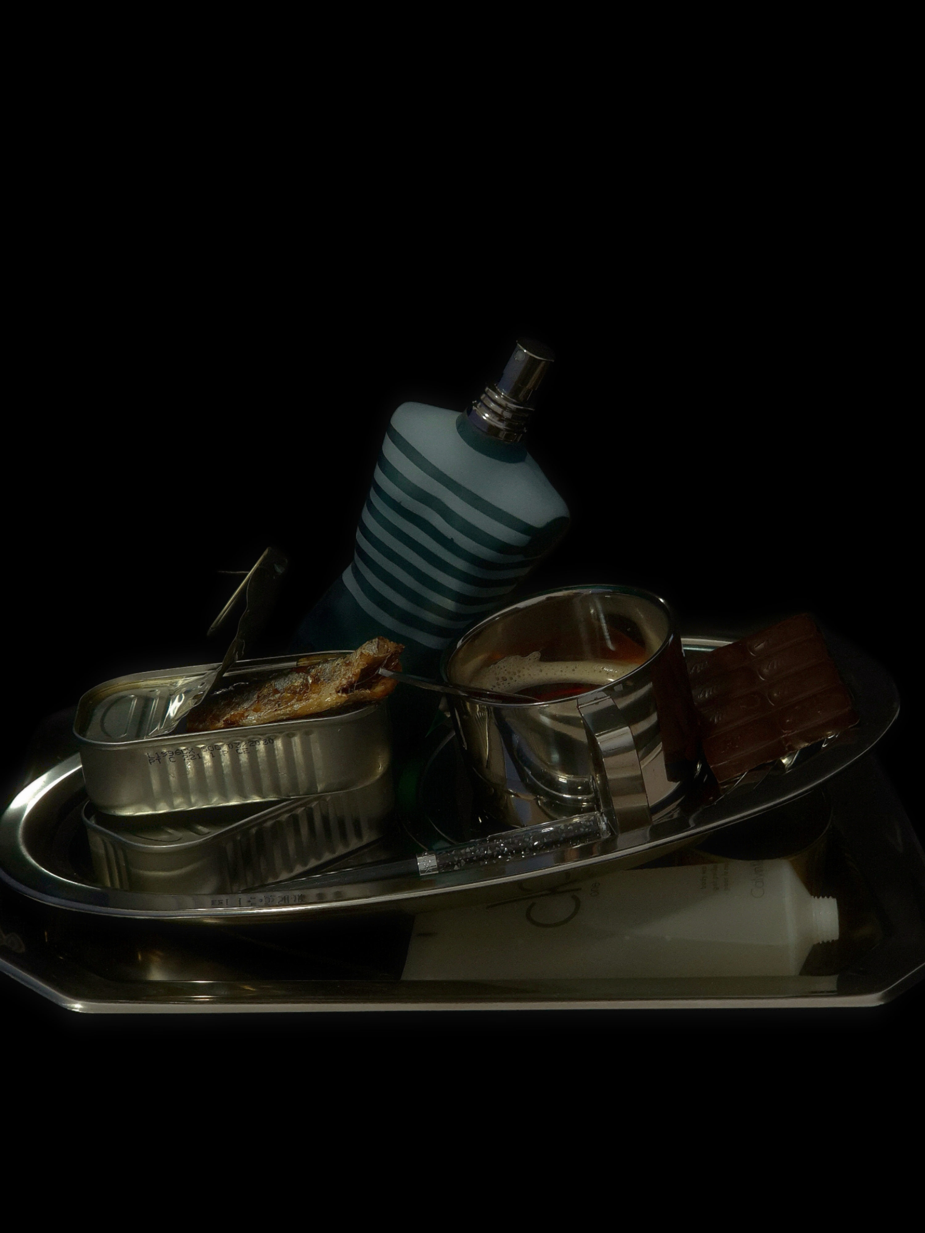 Still life of a perfume bottle, a tin of sardines, a chocolate bar, a metal cup, and a pen arranged together on a dark reflective surface.