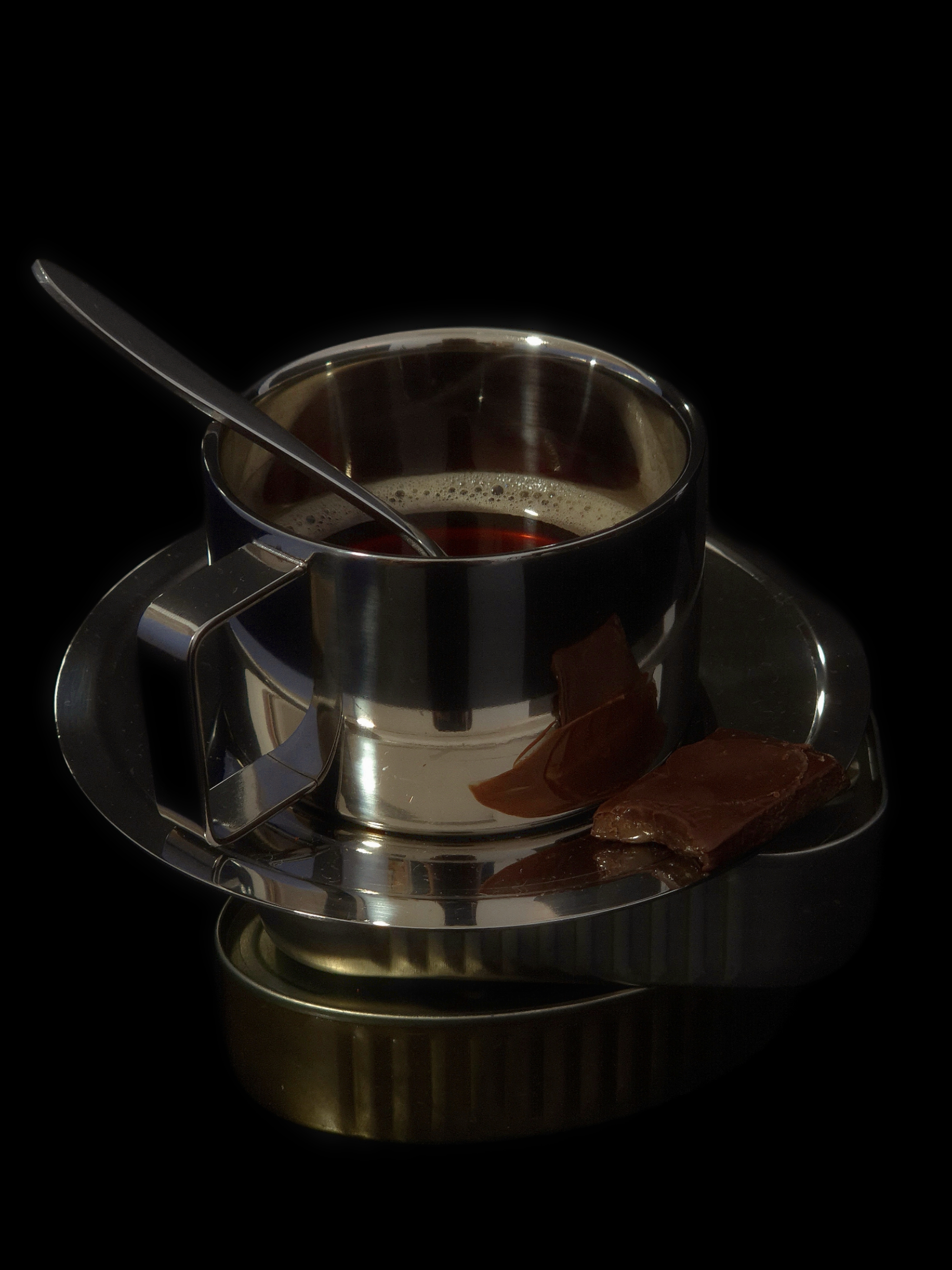 Still life of a metal cup placed on a tin, with pieces of chocolate beside it against a dark background.
