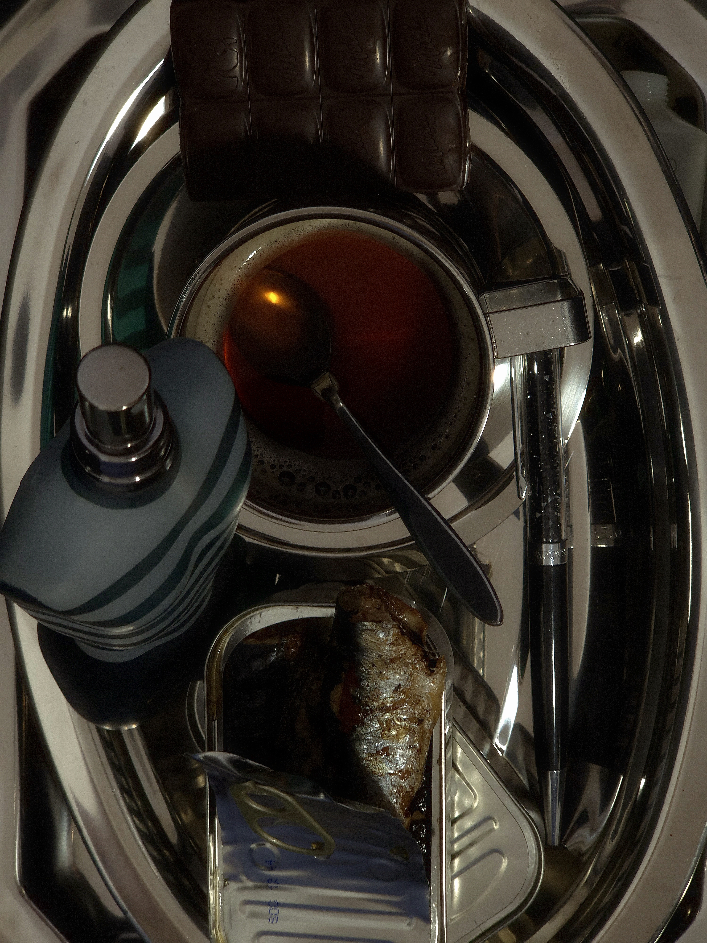 Top-view still life showing a metal cup, a chocolate bar, a perfume bottle, and a tin of sardines arranged on a dark surface.

