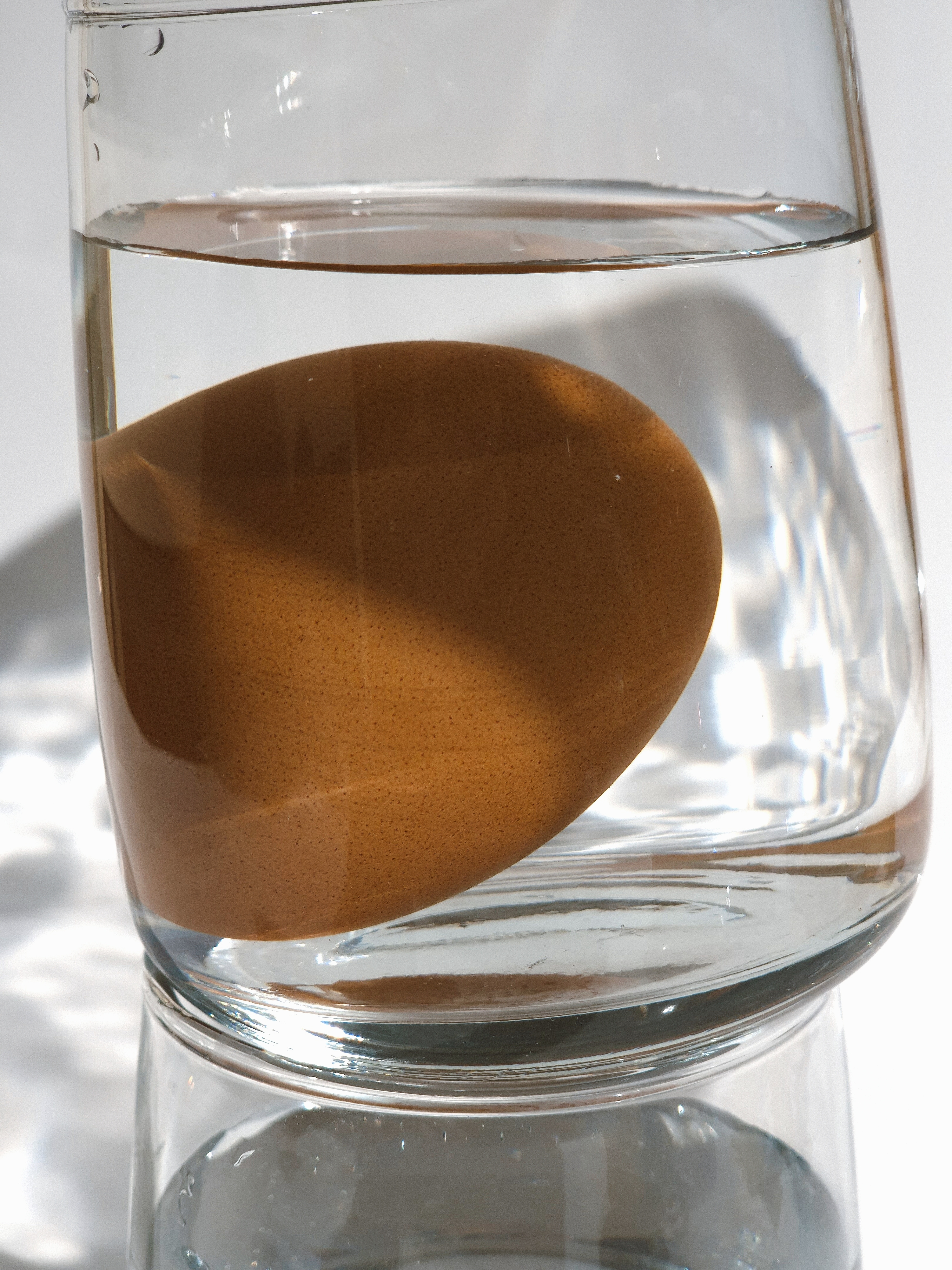 Egg suspended in water inside a tilted glass under direct light.