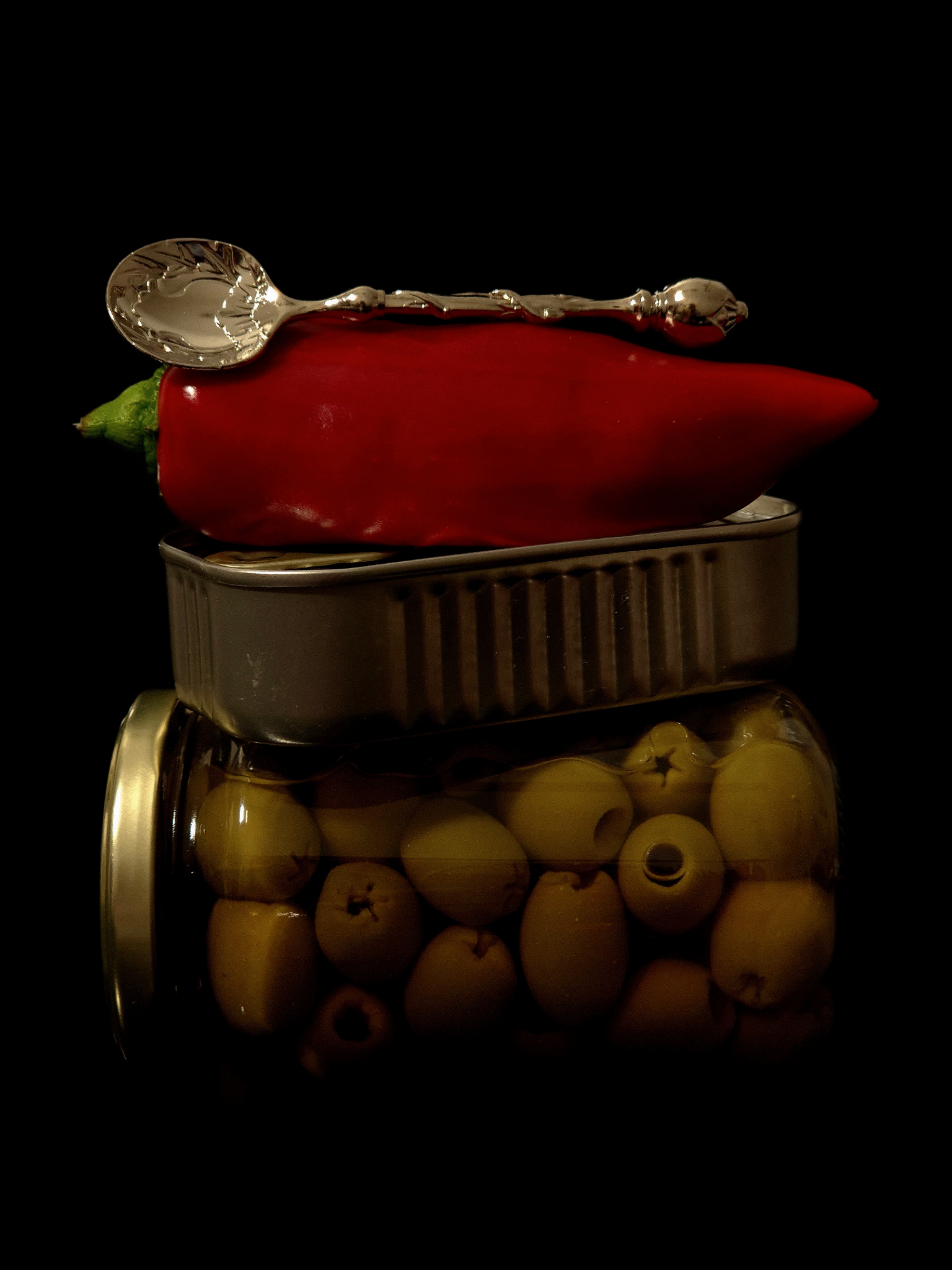 Gourmet still life with a red pepper topped by a silver spoon, placed on a tin above a jar of olives against a dark background.