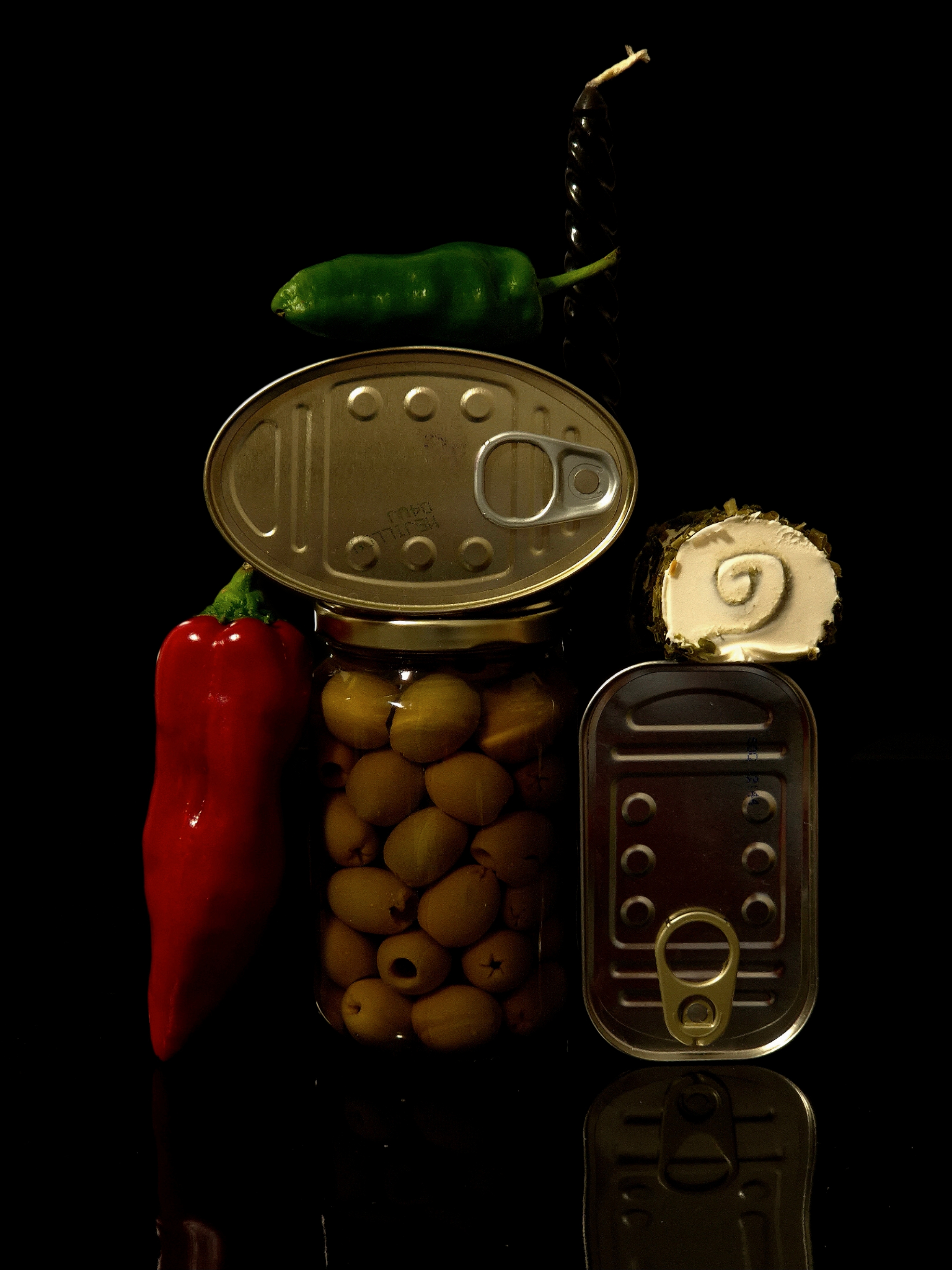Gourmet still life with a jar of olives, two tin can lids, a red pepper, a green pepper, and a piece of cheese arranged against a dark background.