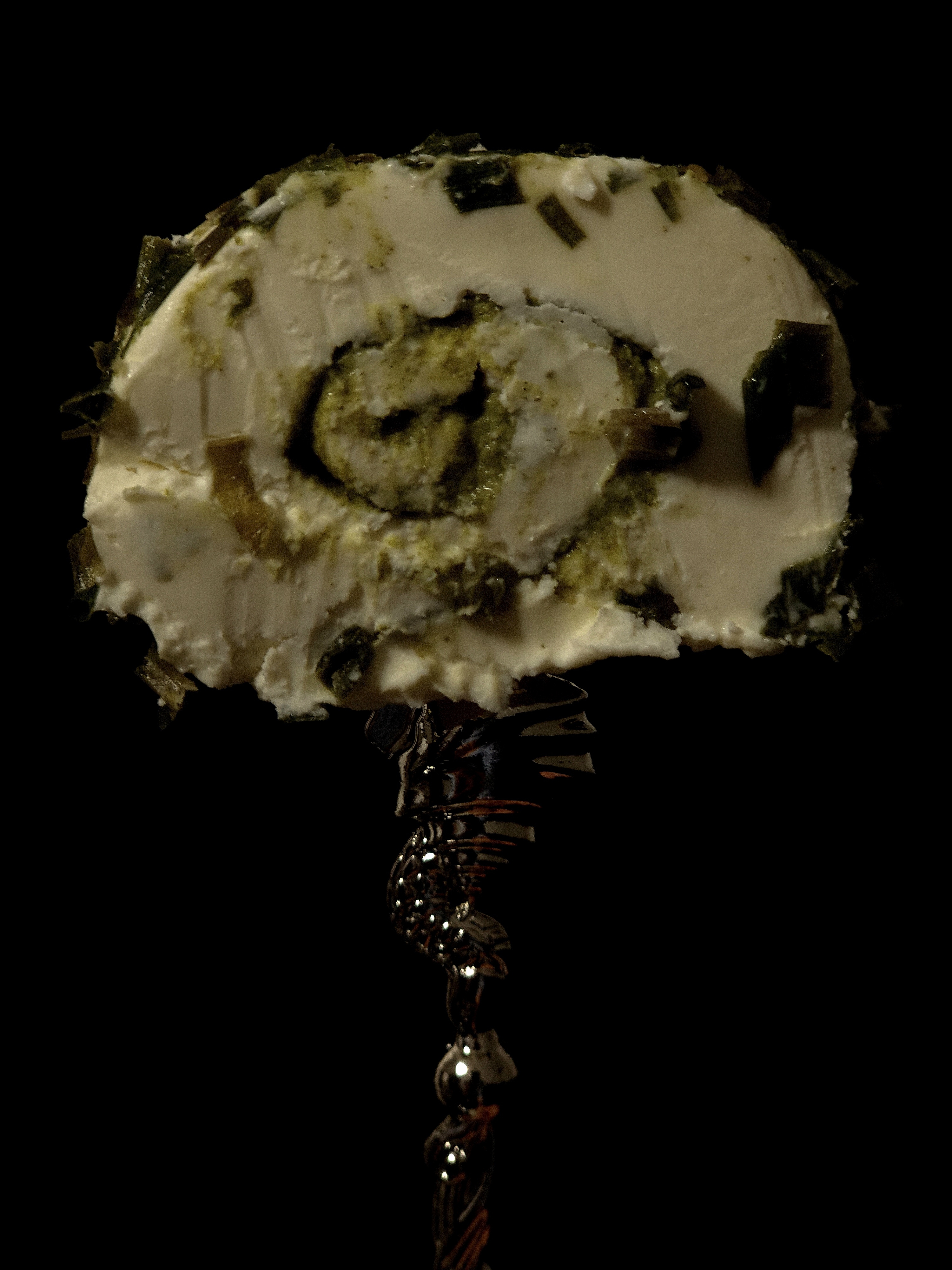 Close-up gourmet still life of a slice of herbed cheese held on an ornate silver utensil against a dark background.