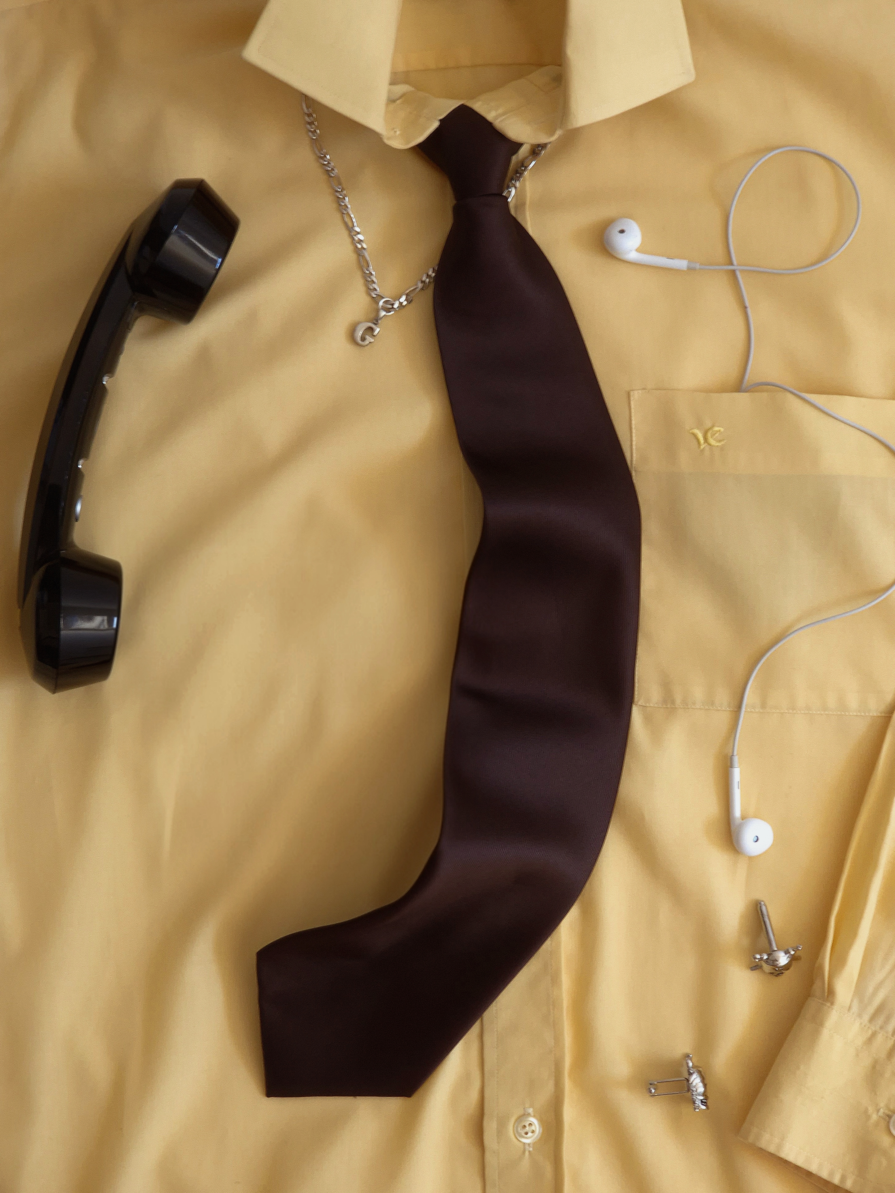Yellow shirt with brown tie styled as a flat lay, with a black phone receiver, earphones and cufflinks.