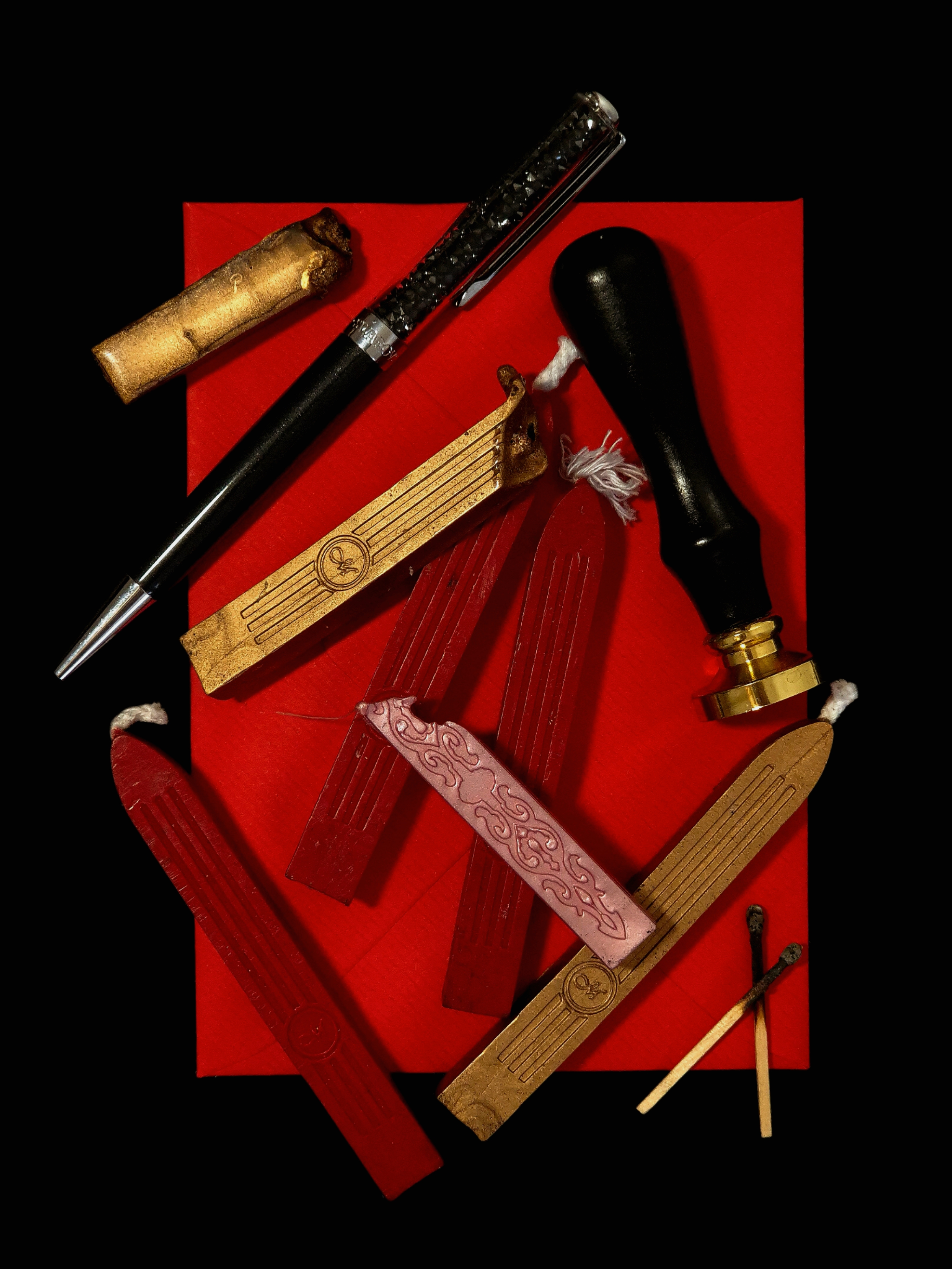 Still life of red wax sticks, a sealing stamp, a black writing instrument and fire-marked matches arranged on a red envelope against a dark background.