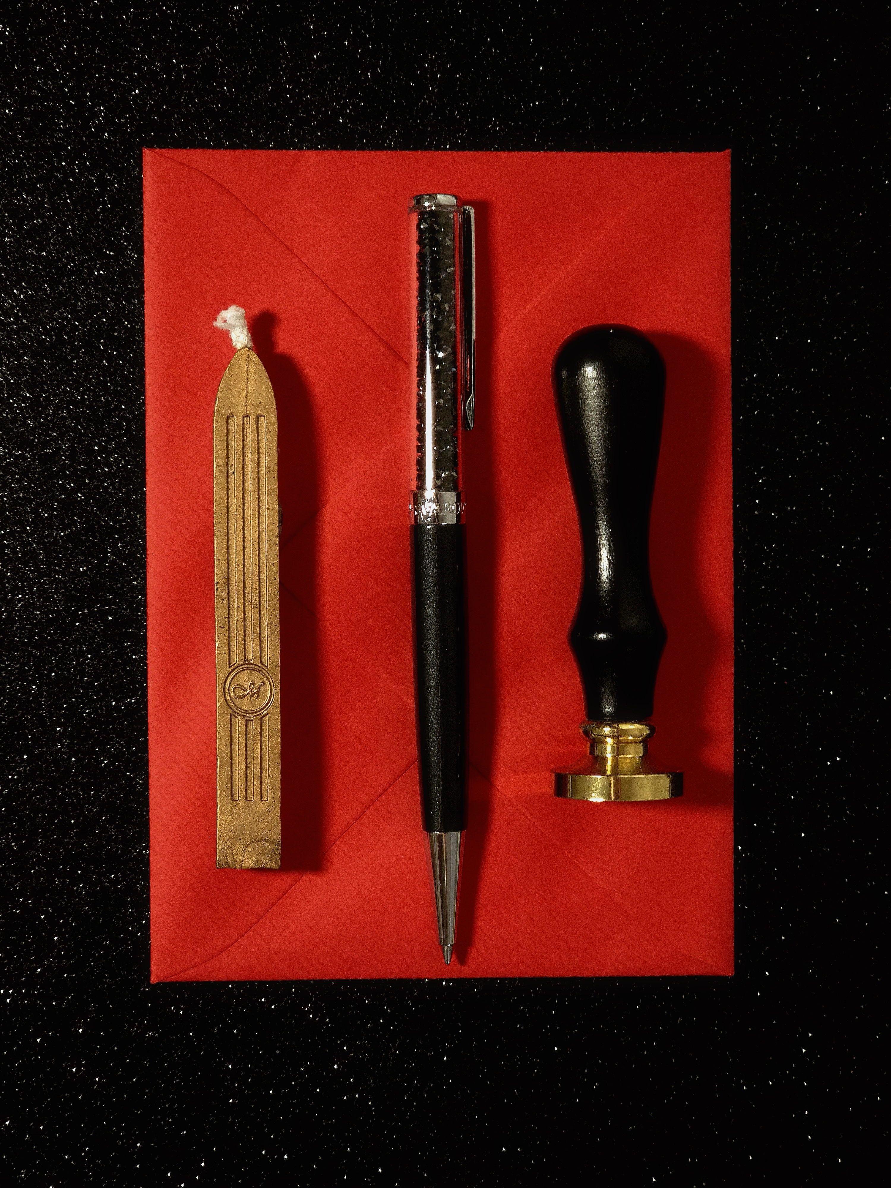 Still life with a gold sealing stamp, a black pen and a red wax stick placed neatly on a red envelope over a dark surface.