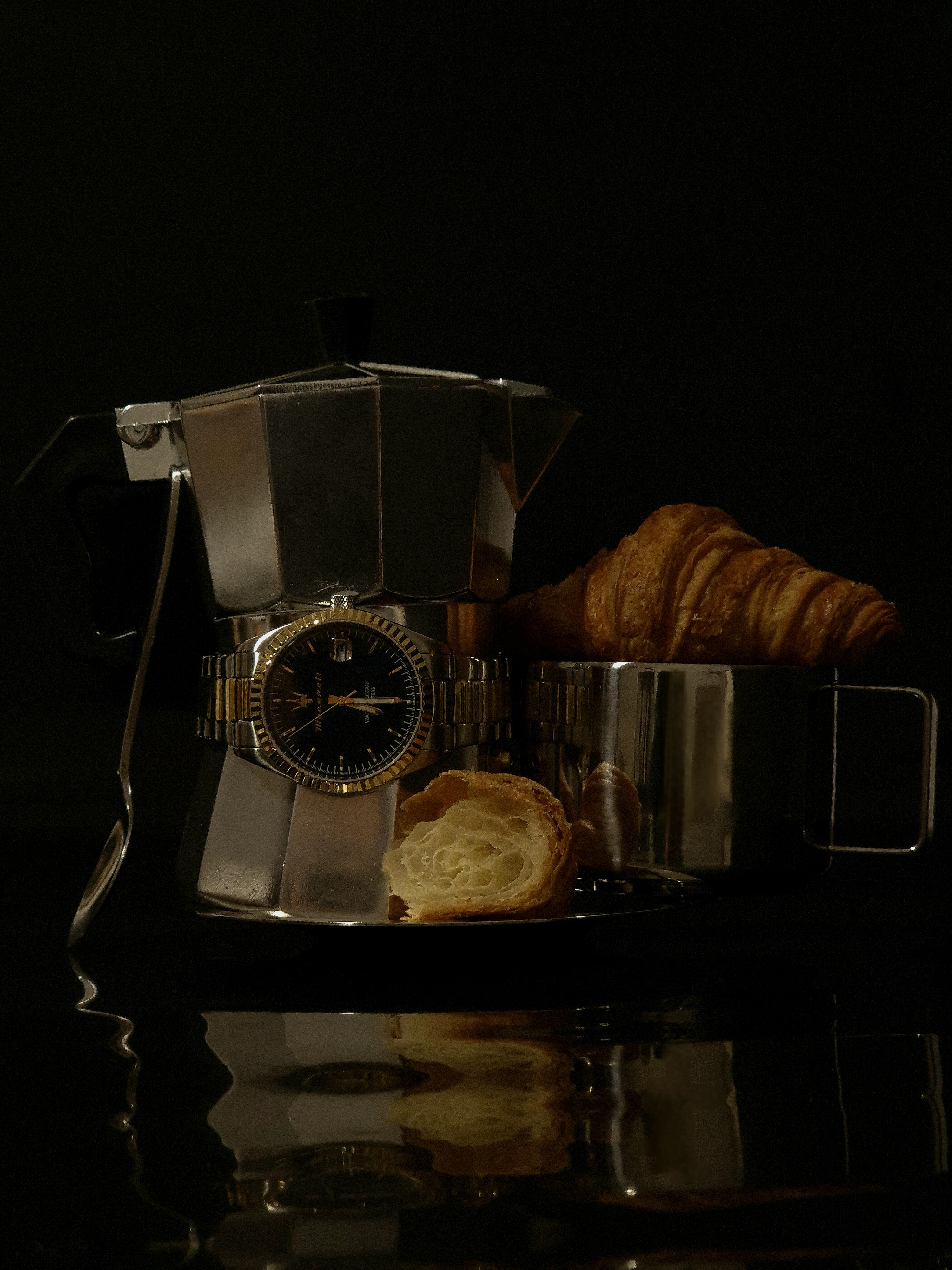 Still life with a moka pot, a stainless-steel cup, croissants and a wristwatch arranged on a reflective black surface.