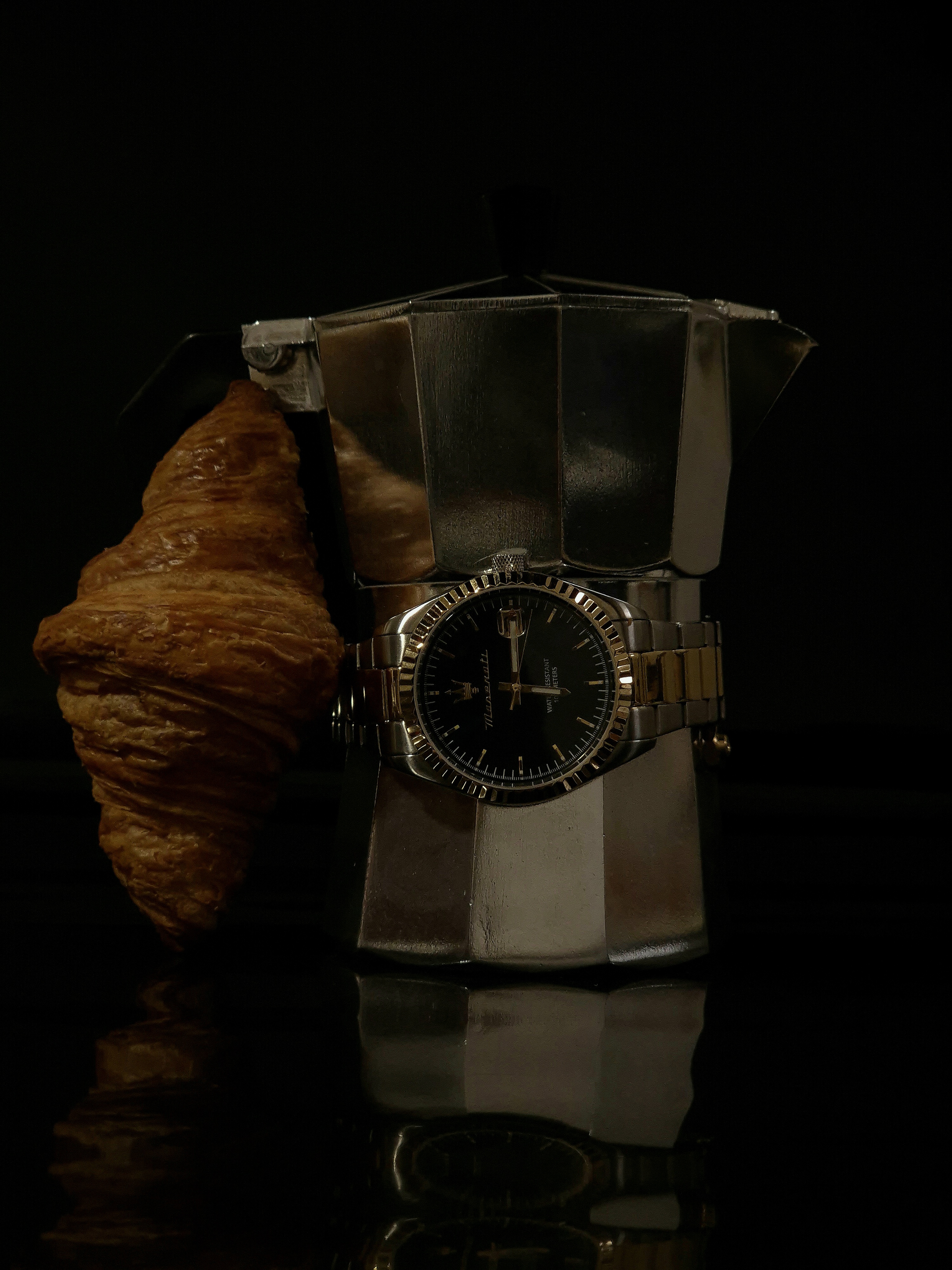 Still life featuring a moka pot with a croissant resting on the handle and a wristwatch draped around the base.
