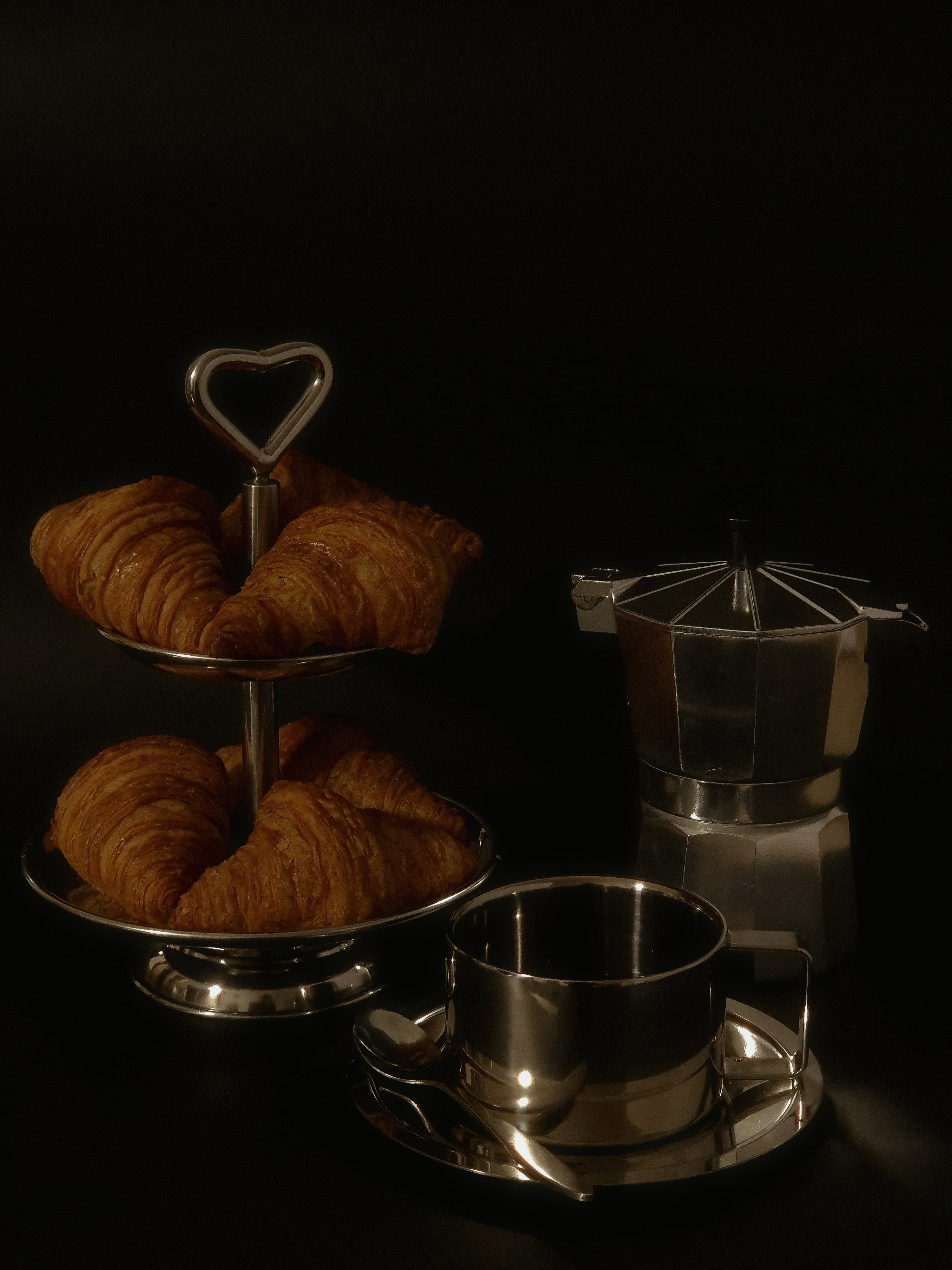 Still life with a metal tiered tray of croissants, a stainless-steel coffee cup, and a moka pot against a dark backdrop.