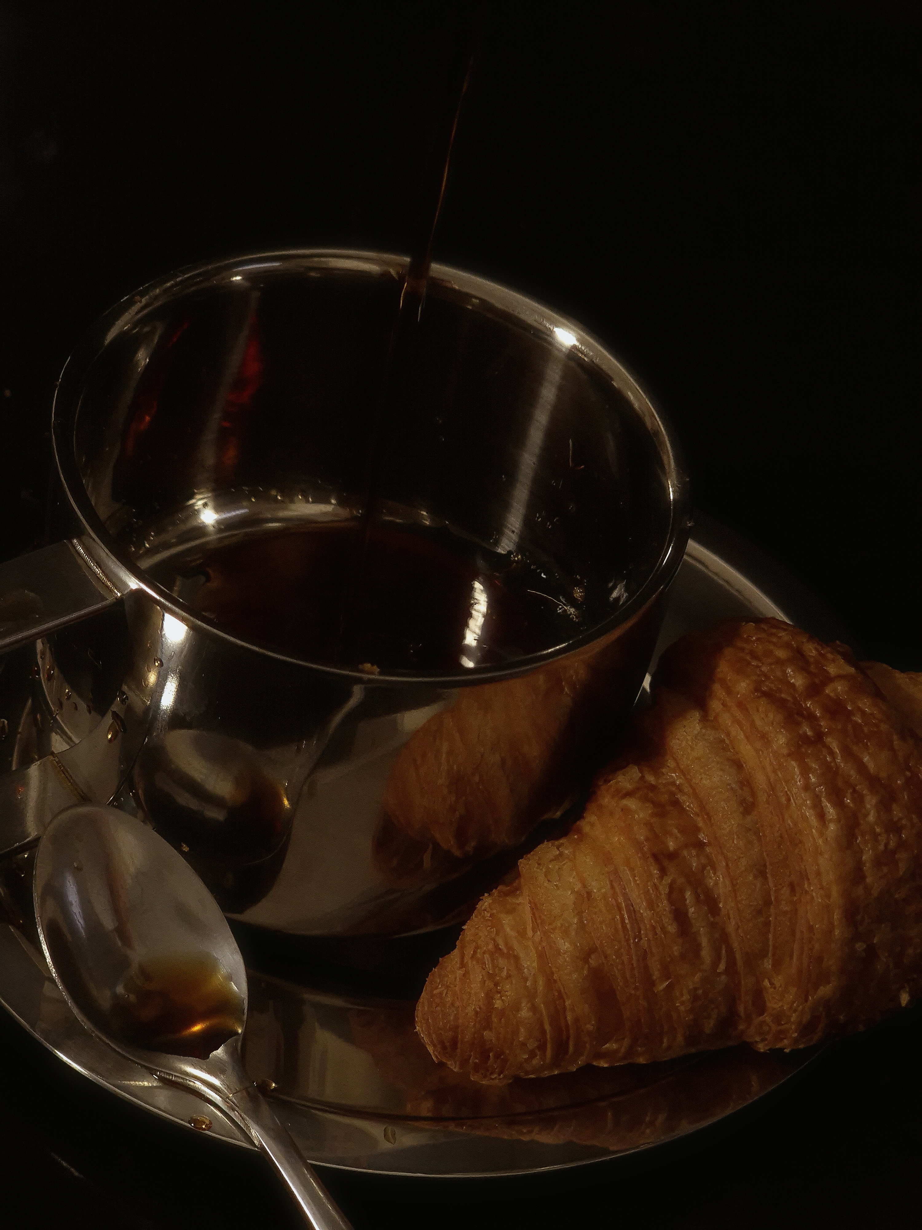 Close-up of coffee being poured into a stainless-steel cup beside a croissant on a reflective surface.