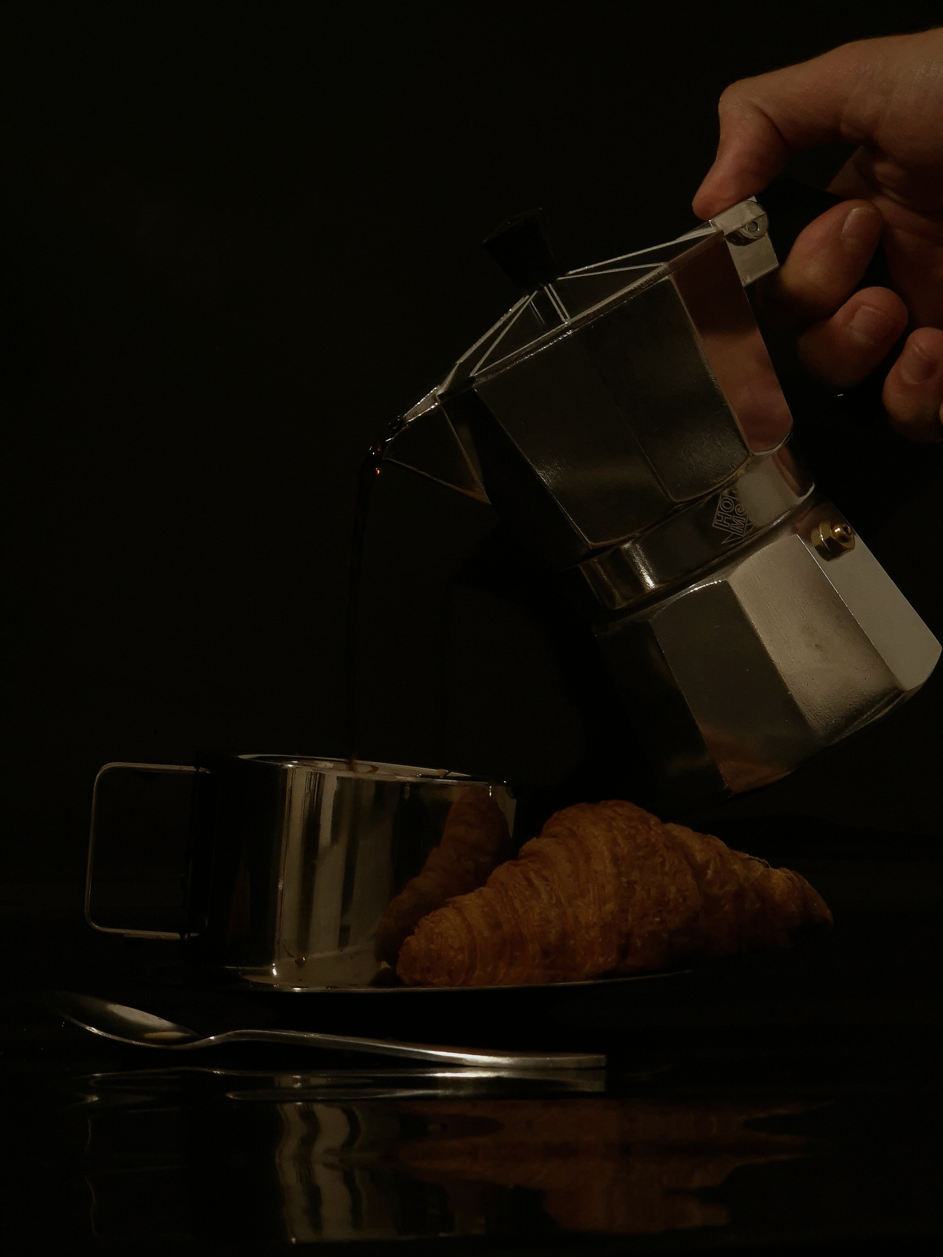 Still life of a hand pouring coffee from a moka pot into a stainless-steel cup next to a croissant.