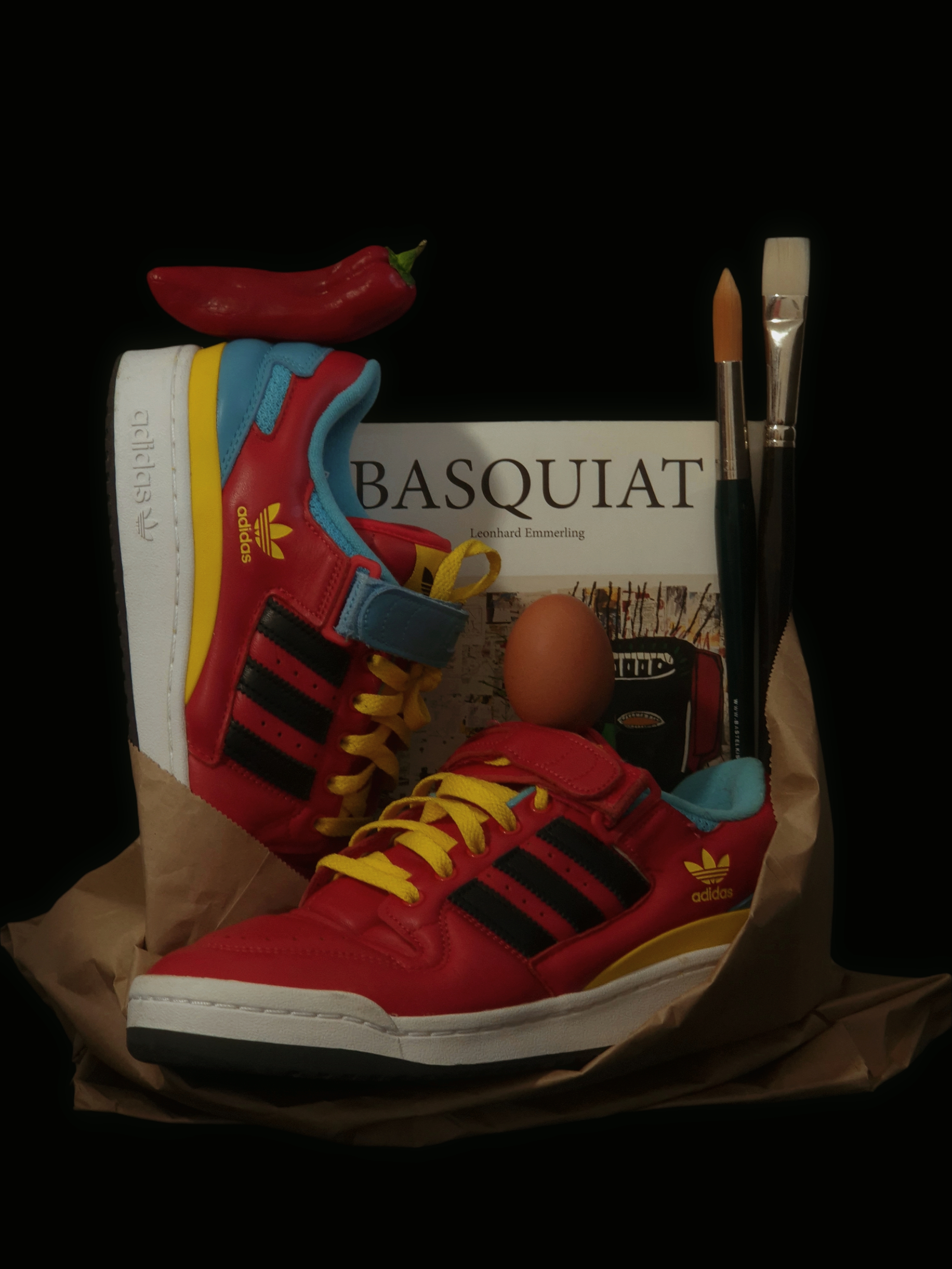Colorful Adidas sneakers styled with a Basquiat art book, a chili pepper, an egg and paintbrushes, arranged as a conceptual still life against a dark background.