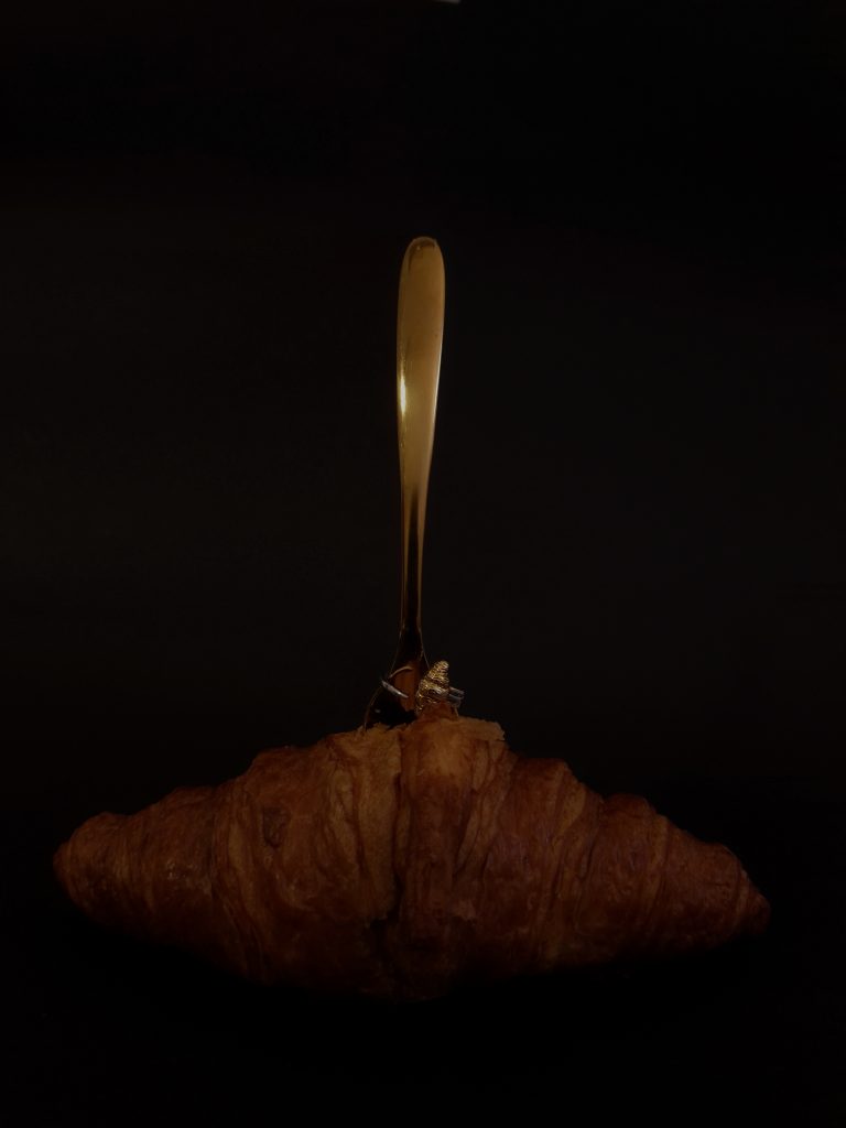 Golden croissant-shaped ring displayed on a fork resting over a croissant, set against a dark background.
 