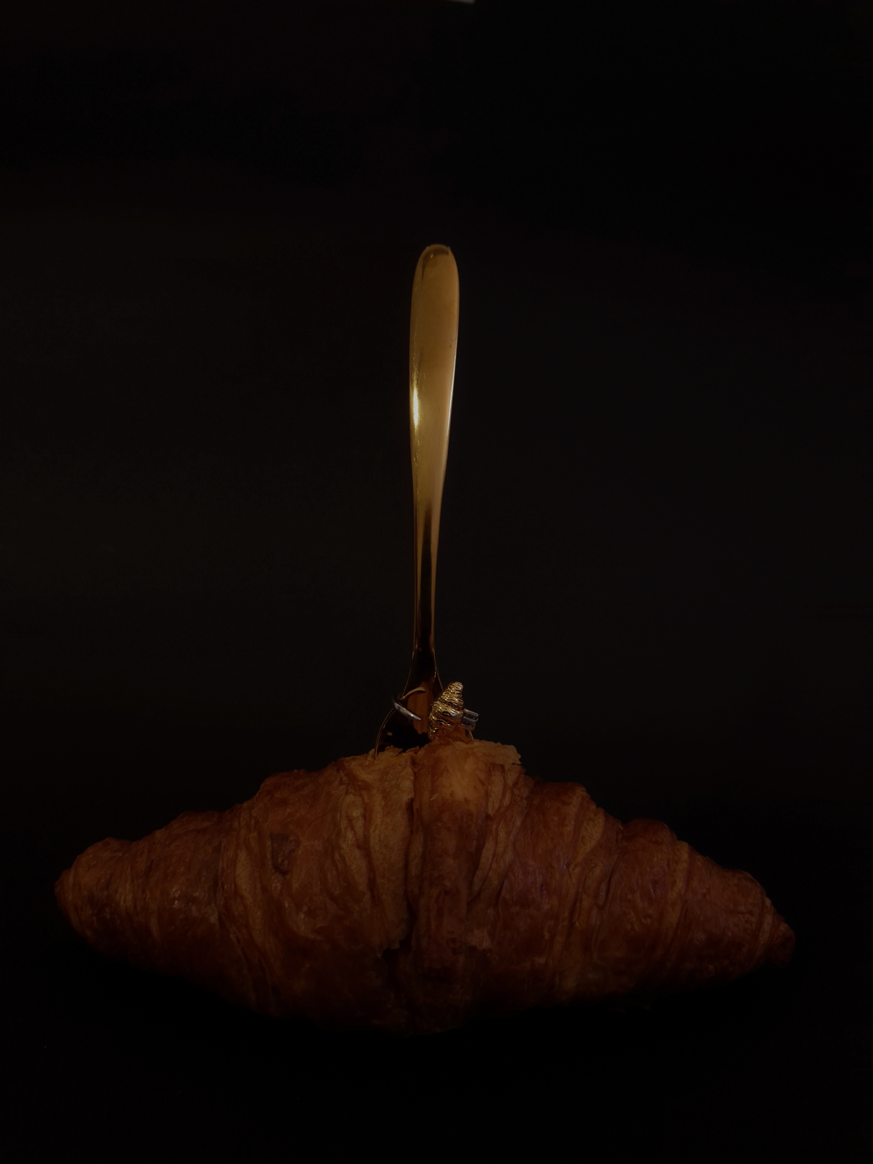 Golden croissant-shaped ring displayed on a fork resting over a croissant, set against a dark background.