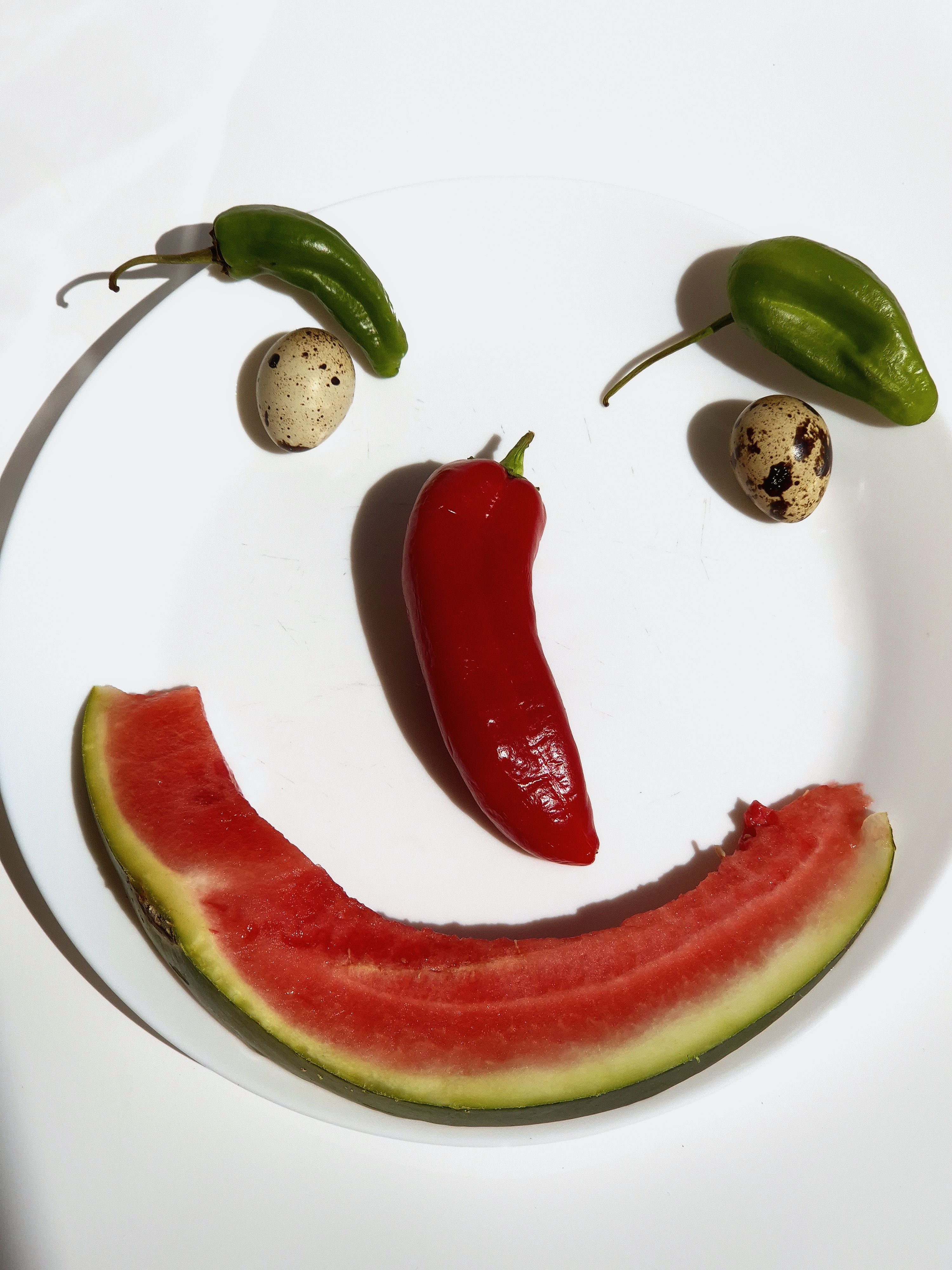 A playful still life arranged to resemble a smiling face, featuring a watermelon slice, peppers, and quail eggs on a white plate, photographed in bright light.
